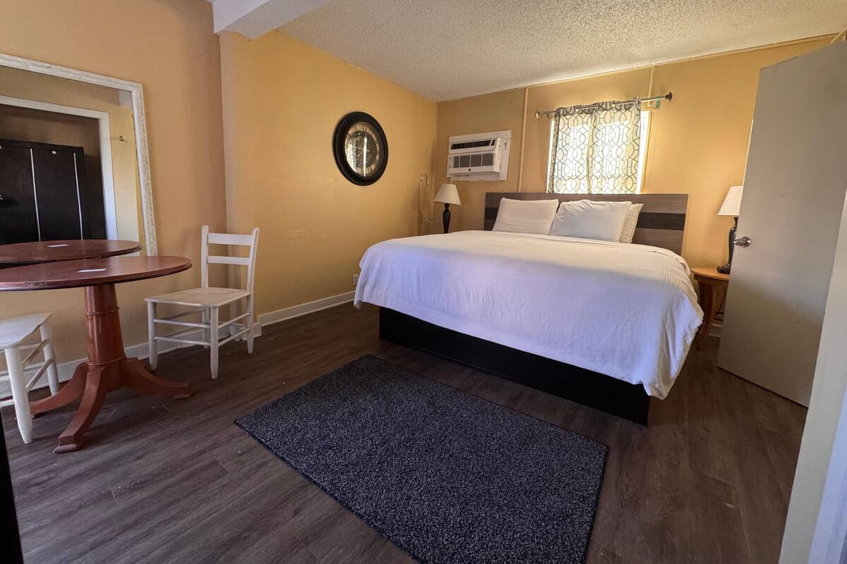 Un dormitorio con cama, escritorio, silla, reloj, equipo de aire acondicionado y una puerta.