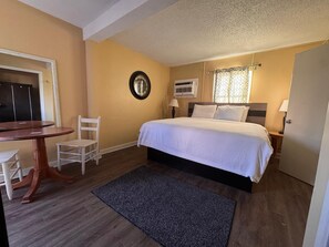 A bedroom with a bed, a desk, a chair, a clock, an air conditioning unit, and a door. - CMI Hotel 1935 Myrtle Beach SC (Myrtle Beach)
