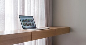 A laptop on a wooden desk with a white mouse, positioned in front of a window with sheer curtains. - Faria Lima by Conviva (São Paulo)