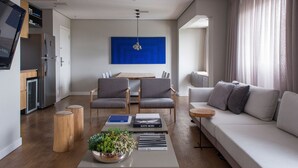 Un salon moderne comprenant un canapé, deux fauteuils, une table basse avec des livres et une plante, un coin repas avec une table et des chaises, et une cuisine équipée d’appareils électroménagers.