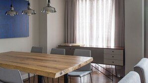 Un espace repas moderne avec une table en bois, des chaises grises, des luminaires suspendus et une vue sur la cuisine à travers une fenêtre ornée de rideaux.