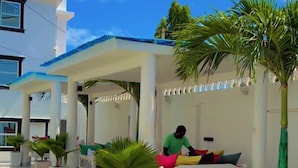 A covered outdoor seating area with a person sitting on a bench, surrounded by potted plants and a palm tree.