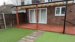 A backyard with artificial grass, a wooden deck, and a brick wall. - Lovely 6-bed House in Windsor (Windsor)