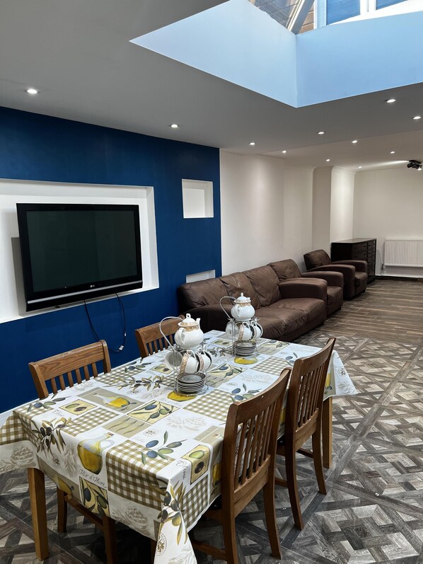 A dining area with a table set for four, a brown sofa, and a television mounted on a blue wall. - Lovely 6-bed House in Windsor (Windsor)