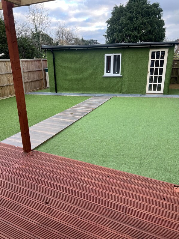 A backyard with artificial green turf, a wooden deck, and a small building with a window and door.