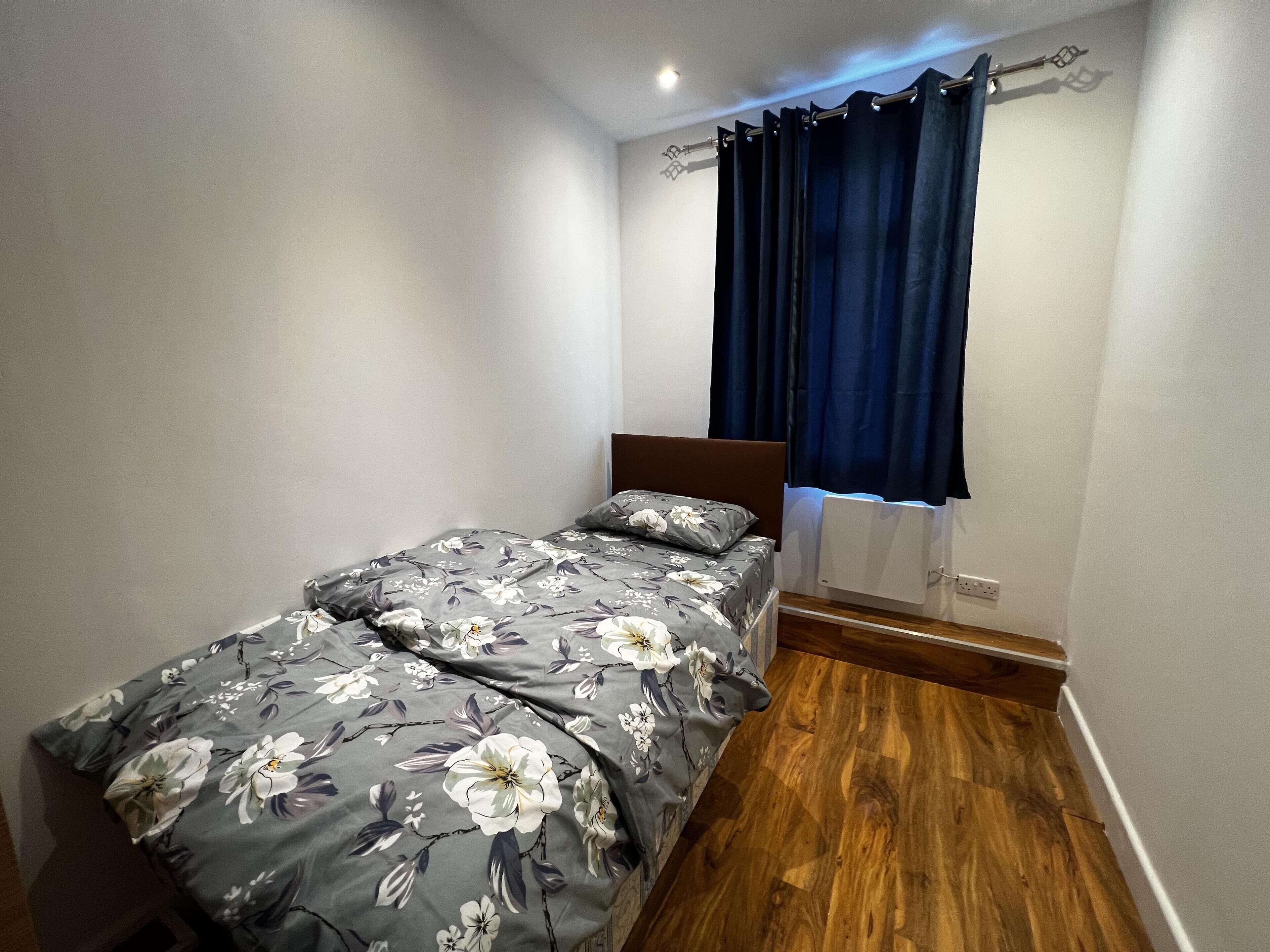 A small, single-bed room with a wooden floor, a dark headboard, and a window with dark curtains.