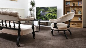 A living room with a sofa, armchair, and a television displaying a scenic landscape. - Cozy 4-bed Cottage Garden Walk to River Thames (Hampton)