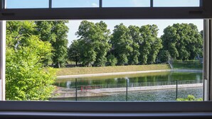 A view through a window of a park with a pond, trees, and a fence. - Cozy 4-bed Cottage Garden Walk to River Thames (Hampton)