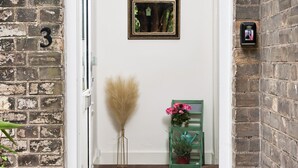 A narrow hallway with a white door, a brick wall, a framed picture on the wall, a small green plant stand, and a potted plant. - Cozy 4-bed Cottage Garden Walk to River Thames (Hampton)