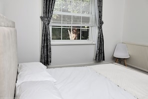 A bedroom with a single bed, a window with curtains, a radiator, and a small table with a lamp.