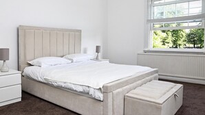 A bedroom with a bed, bedside tables, a window with a view of trees, and a radiator. - Cozy 4-bed Cottage Garden Walk to River Thames (Hampton)
