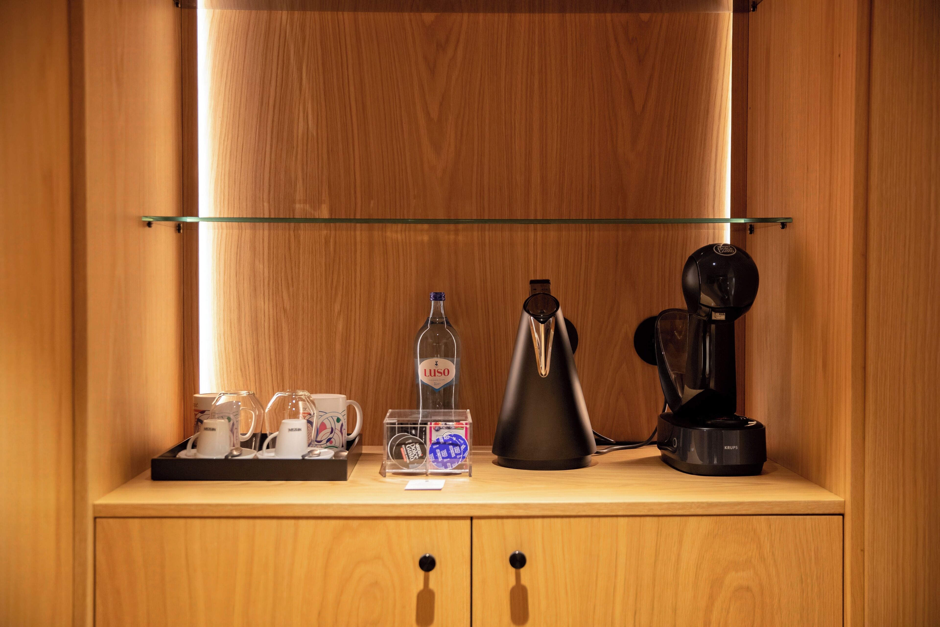 Un mueble de madera con estante de vidrio que contiene una cafetera, una botella de Luso y una bandeja con tazas y una tetera.