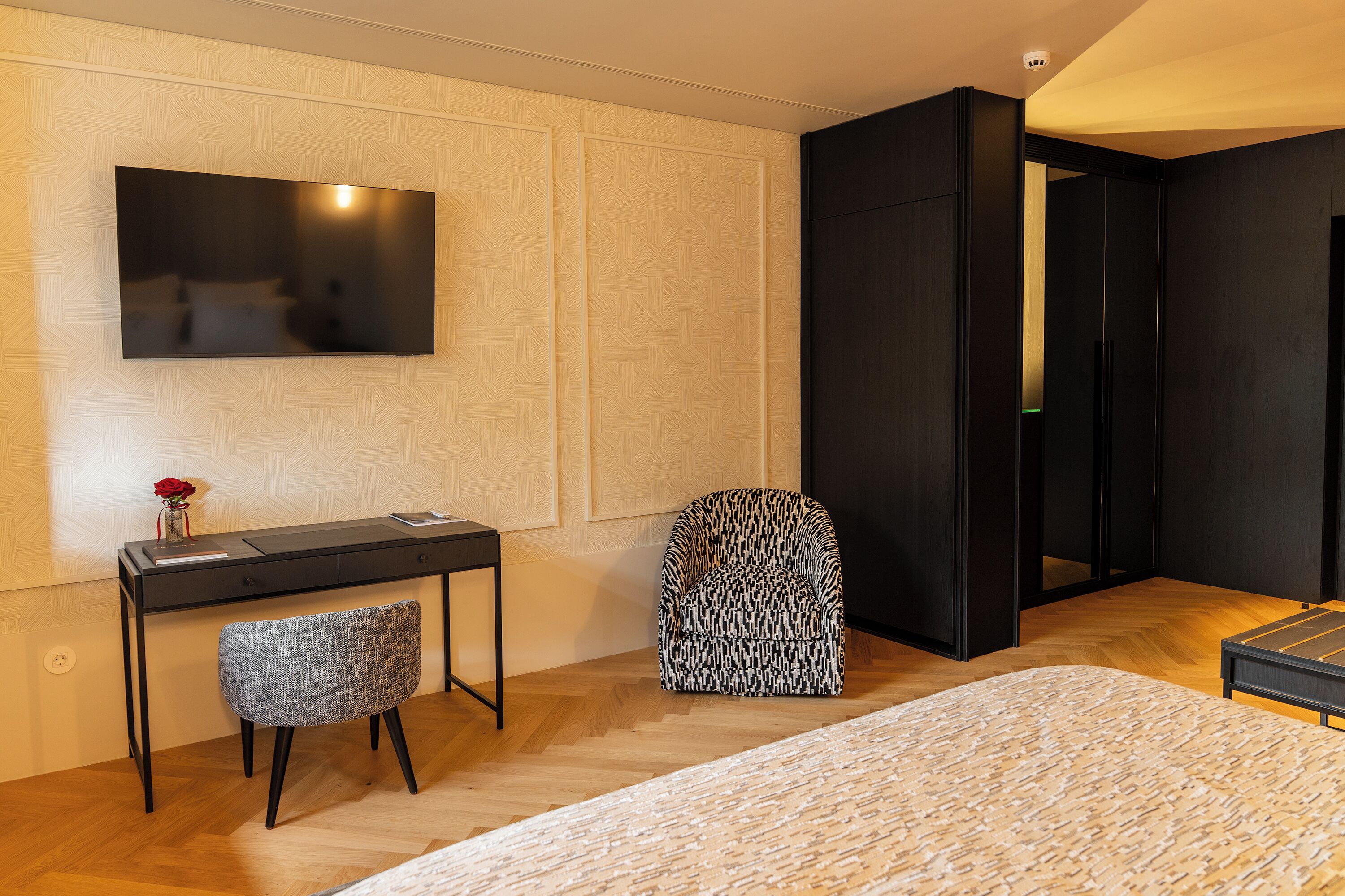 A modern hotel room with a bed, a desk with a chair, a television mounted on the wall, and a wardrobe.
