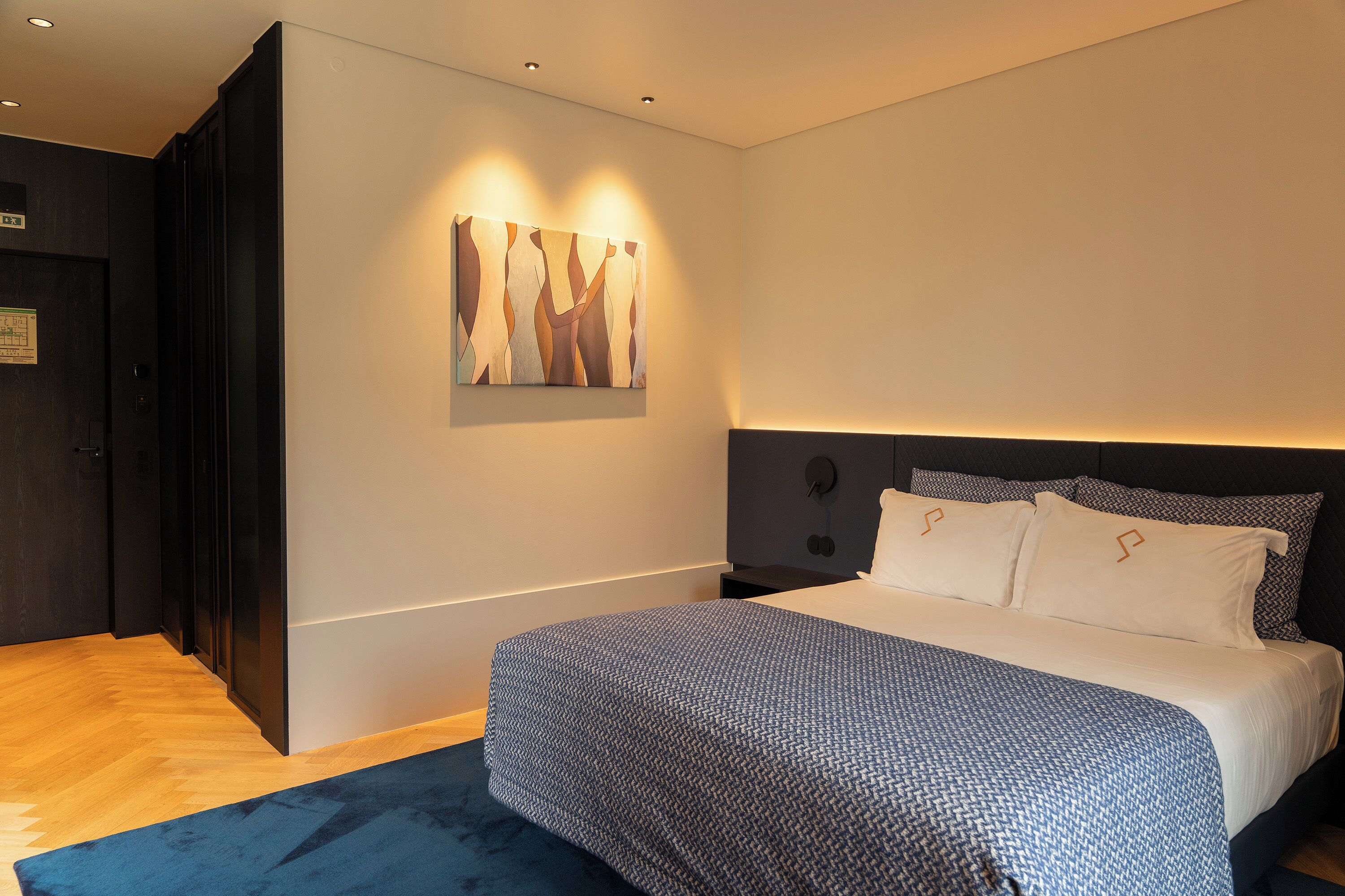 Una habitación de hotel moderna con una cama grande, cabecera con iluminación integrada, una obra de arte colgada en la pared y una puerta de madera oscura.