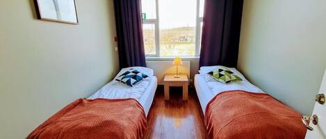 Una habitación con dos camas, una mesita con lámpara, una ventana con cortinas y un cuadro en la pared.