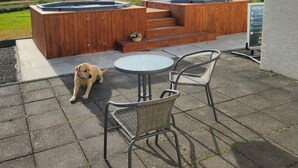 Un bain à remous en bois avec des marches, deux chaises en métal et un chien sur une terrasse offrant une vue sur les montagnes lointaines.