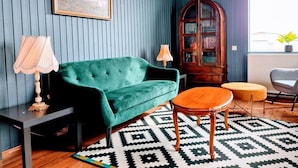 Une pièce avec un canapé en velours vert, une table d’appoint en bois, un tapis à motifs, un meuble en bois et une fenêtre donnant sur un paysage agréable.