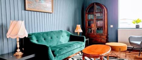 A room with a green velvet sofa, a wooden side table, a patterned rug, a wooden cabinet, and a window with a view.