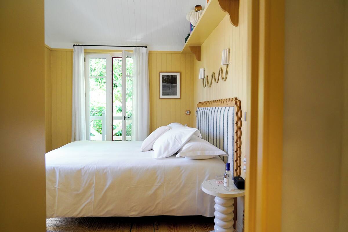 Une chambre à coucher avec un lit, une table de chevet, une fenêtre avec des rideaux, un tableau au mur et une tête de lit en bois.