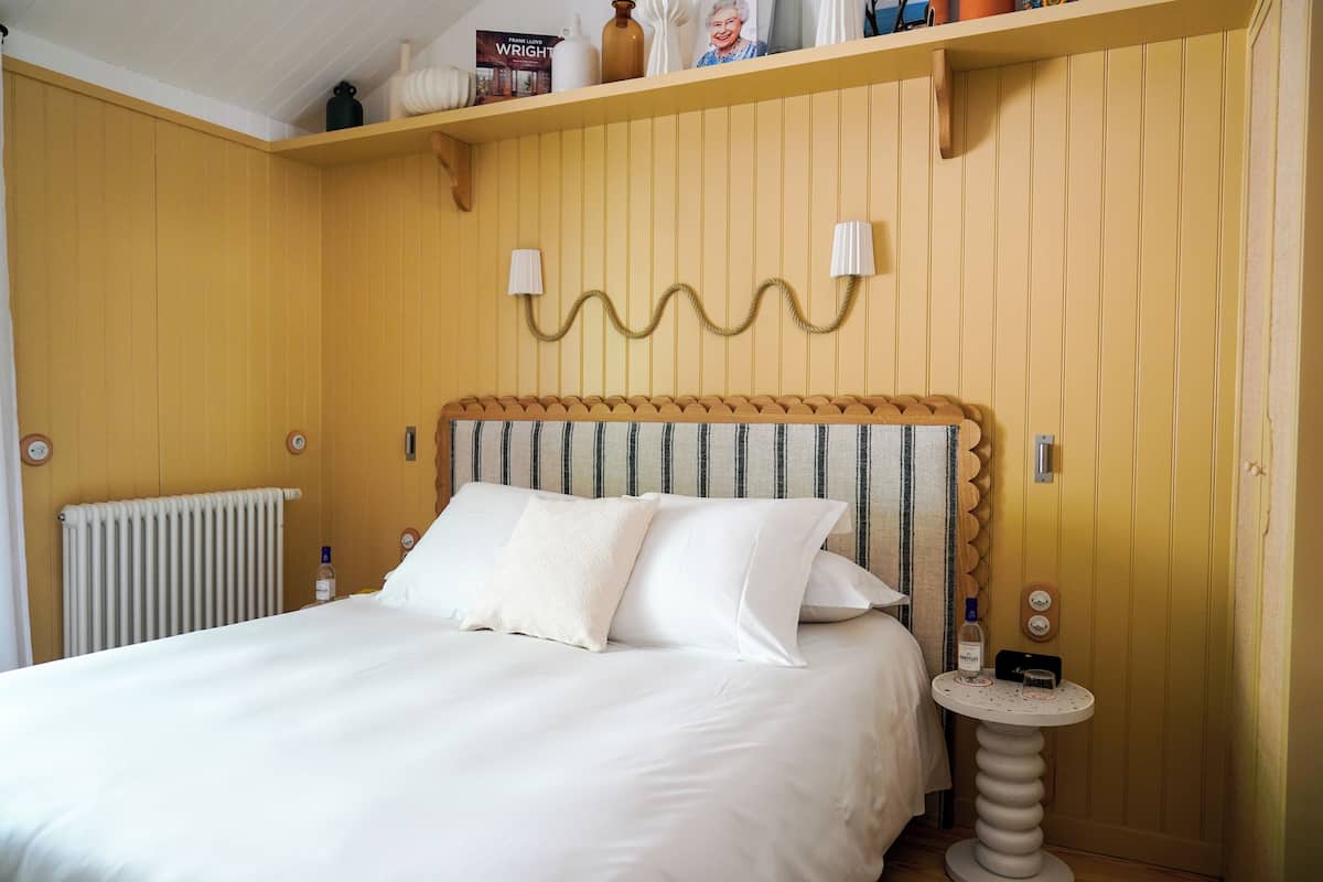 Un lit bien fait, avec du linge de lit blanc et des oreillers, une table de chevet sur laquelle se trouve une bouteille, et une lampe fixée au mur.