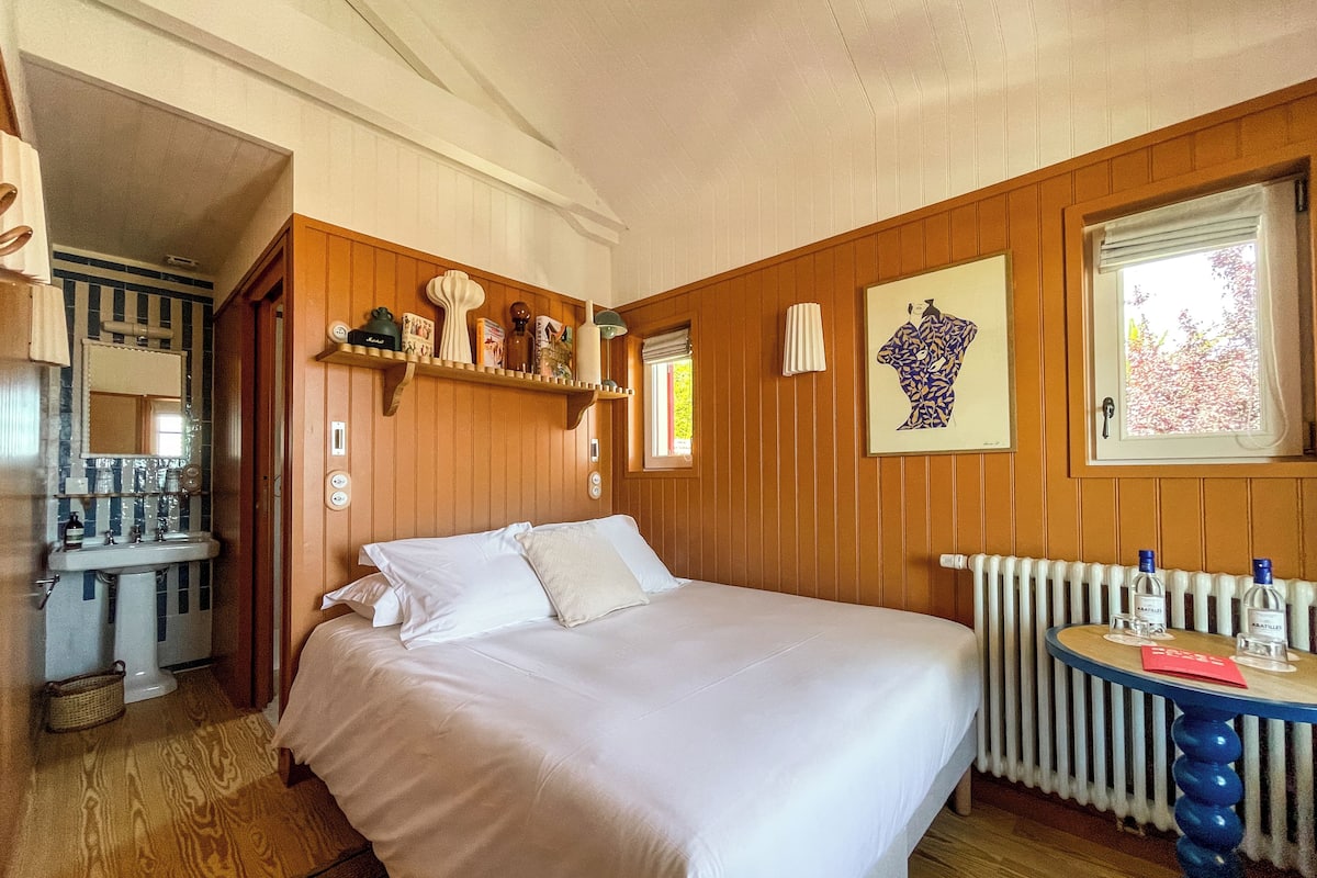 Une chambre à coucher avec une tête de lit en bois, un lit recouvert de draps blancs, une table de chevet sur laquelle reposent deux bouteilles, un radiateur et une œuvre d’art encadrée accrochée au mur.