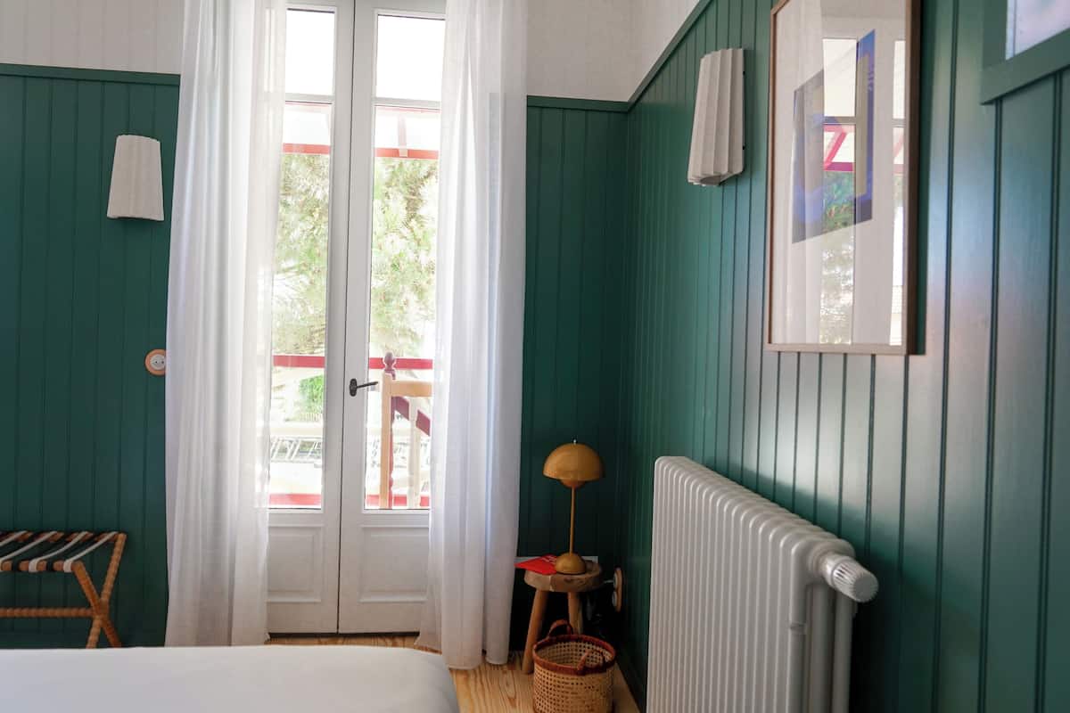 Une pièce aux murs de couleur turquoise, avec un radiateur blanc, un banc en bois, une petite table sur laquelle se trouve une lampe, et une fenêtre avec des rideaux.