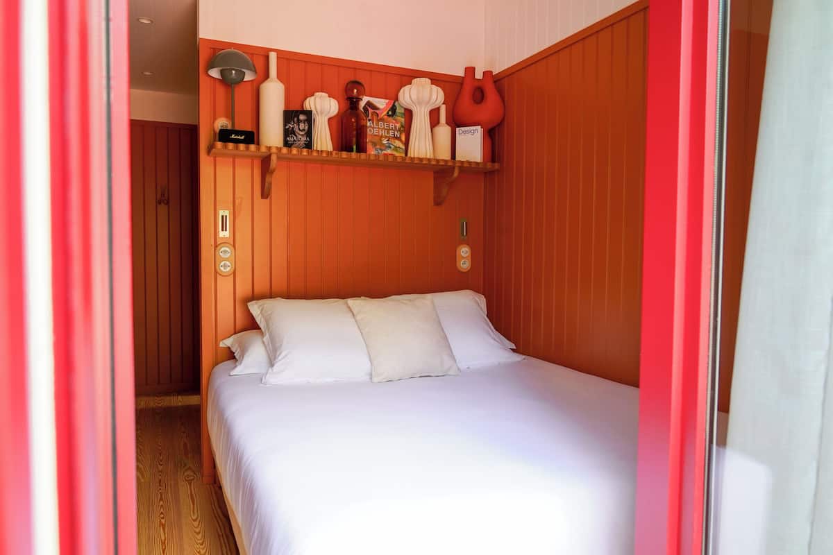 Une chambre d’hôtel avec une tête de lit en bois, un lit recouvert de draps blancs, une étagère ornée d’objets décoratifs et une porte donnant sur une autre pièce.