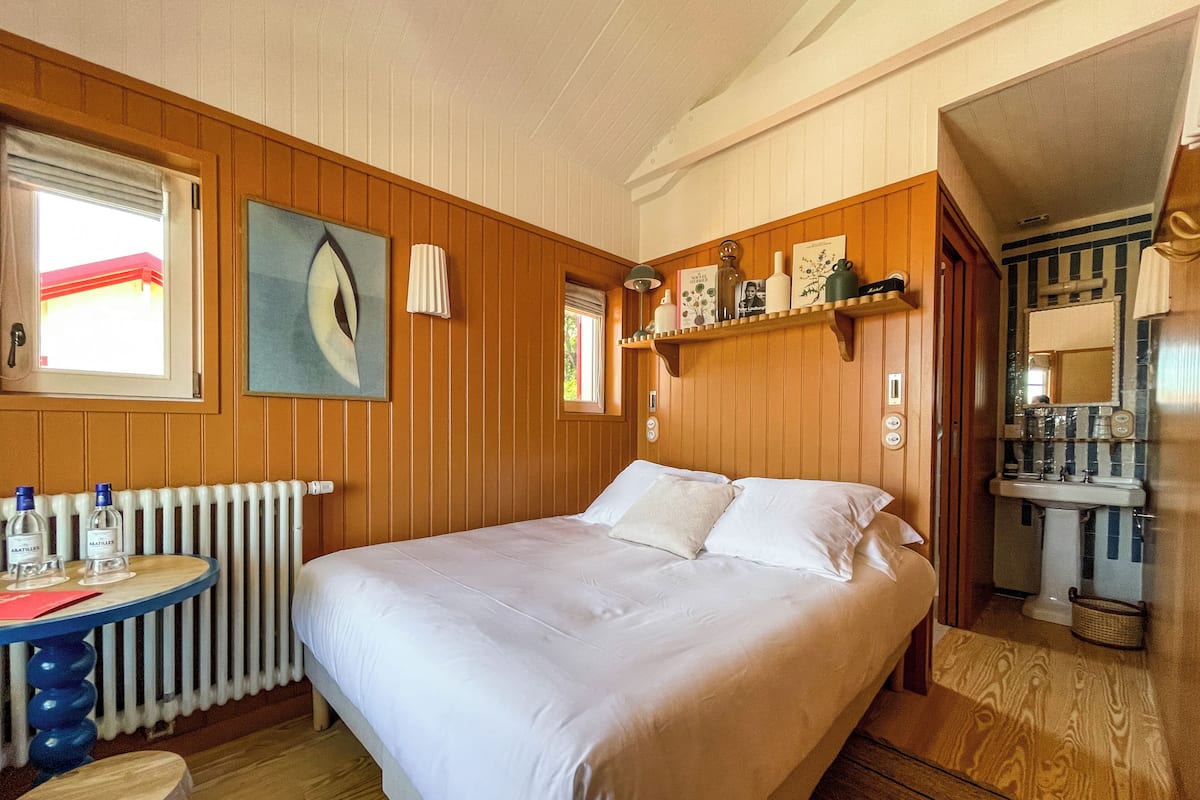 Une chambre avec un grand lit, des murs en bois, un radiateur, une petite table avec des bouteilles et une fenêtre avec des rideaux.