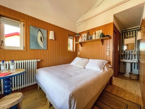 A bedroom with a large bed, wooden walls, a radiator, a small table with bottles, and a window with curtains. - Hôtel de la Plage (Lège-Cap-Ferret)