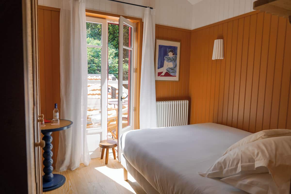 Une chambre à coucher avec un lit, une petite table sur laquelle se trouve une bouteille, un tabouret et un tableau accroché au mur.