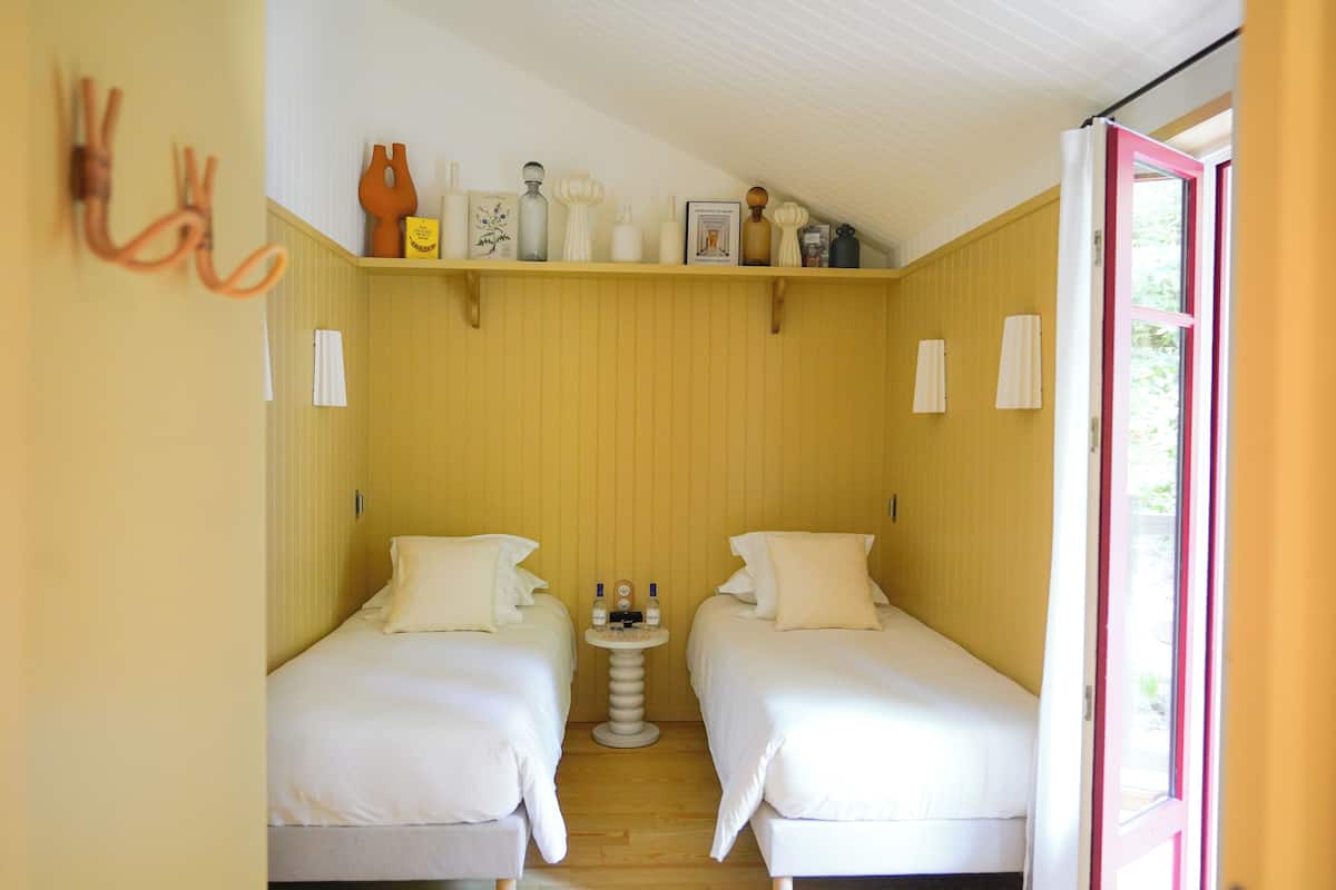 Une petite pièce, de taille modeste, comprenant deux lits simples, une petite table avec des bouteilles et une étagère ornée d’objets décoratifs.