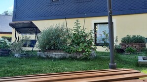 Ein Haus mit dunklem Dach, einer beige farbenen Fassade und einem Garten mit Pflanzen und einer hölzernen Bank.