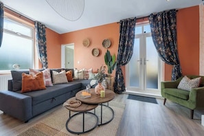 Un salon avec un canapé, une table basse et un fauteuil. On y trouve des rideaux, une plante et un mur décoré d’assiettes.