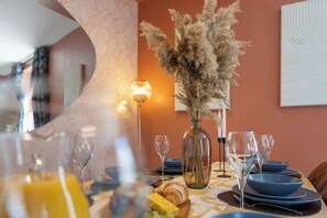 Une table à manger dressée avec de l’assiette bleue, un vase contenant des plantes séchées et une bouteille de jus d’orange.
