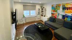 Una sala de estar moderna con un sofá gris, una mesa de centro negra y otra mesa de centro de vidrio. Se ve una estantería con libros y objetos decorativos, una ventana con persianas y un cuadro abstracto y colorido en la pared.