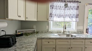 Una cocina con encimera de granito, pileta de acero inoxidable, muebles beige y una ventana con cortinas estampadas.