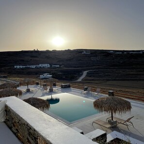 Una piscina en la azotea con sillones de descanso y sombrillas, con vistas a un paisaje montañoso y edificios al fondo.