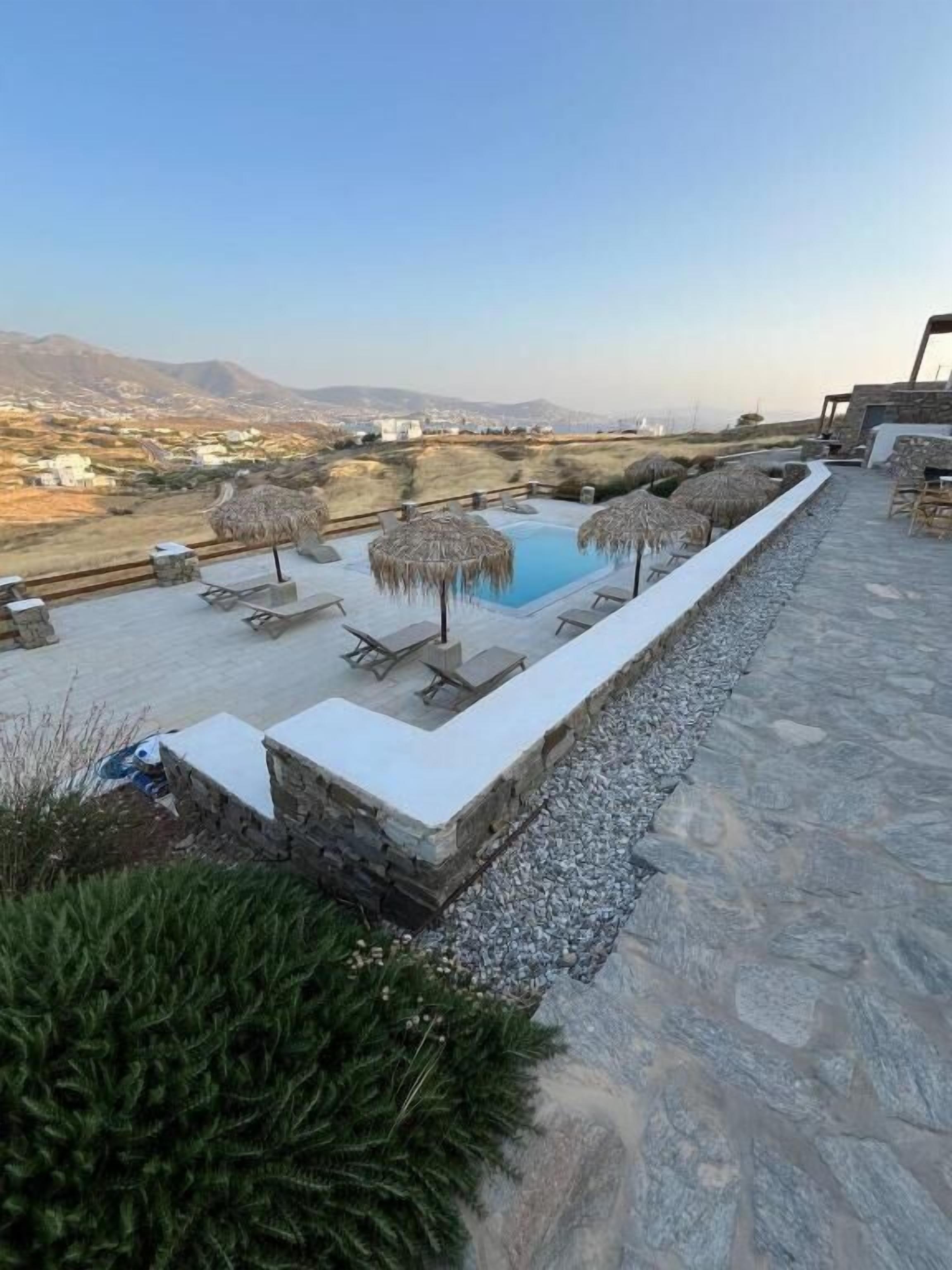 Un espace piscine aménagé sur le toit, avec des chaises longues et des parasols, entouré de murs en pierre et offrant une vue sur les montagnes lointaines.