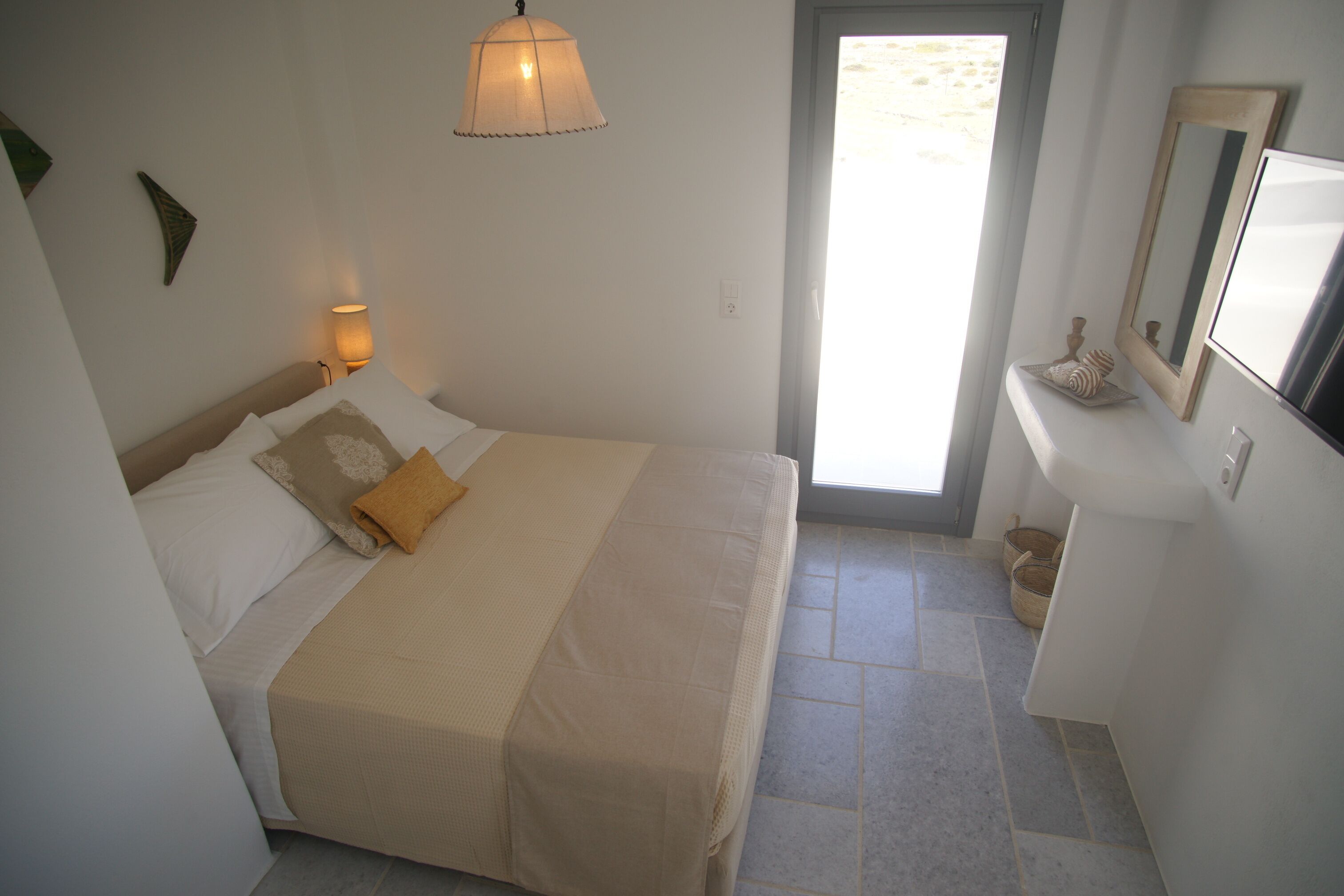 A bedroom with a bed, a bedside lamp, a mirror, and a view of the outside.