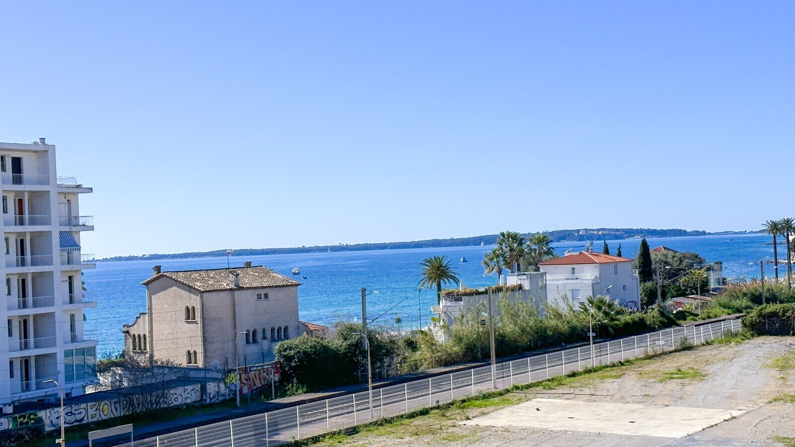 Un bâtiment situé sur la côte, doté d’un balcon, une route bordée d’une clôture et une vue dégagée sur la mer.