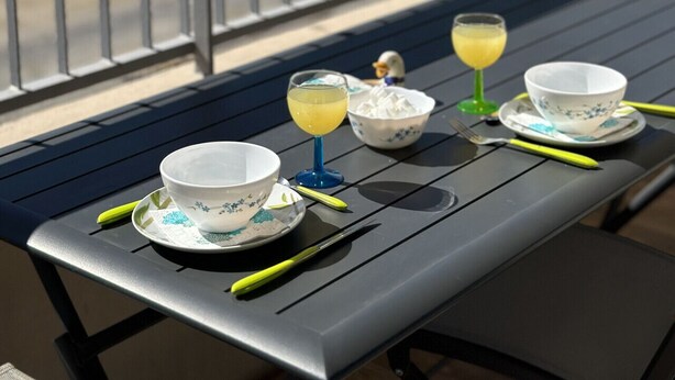 A balcony with a table set for two, featuring a bowl and a glass of drink, overlooking a coastal area.