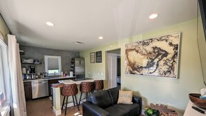 A modern kitchen with a refrigerator, microwave, and coffee maker. There is a dining area with a black leather couch and a coffee table. A large abstract painting is on the wall. - Sleek and Central Small Home ~ Close to everything (Flint)
