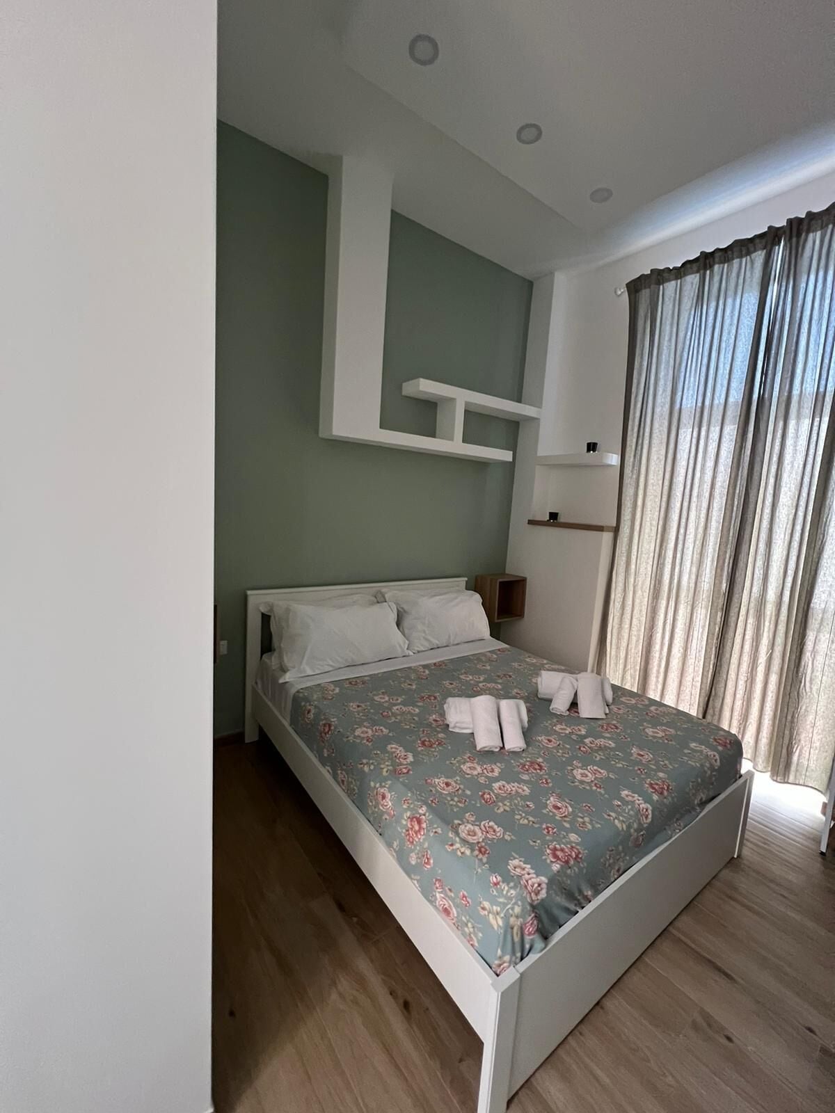 Un dormitorio con cama, mesita de noche, estante en la pared y ventana con cortinas.
