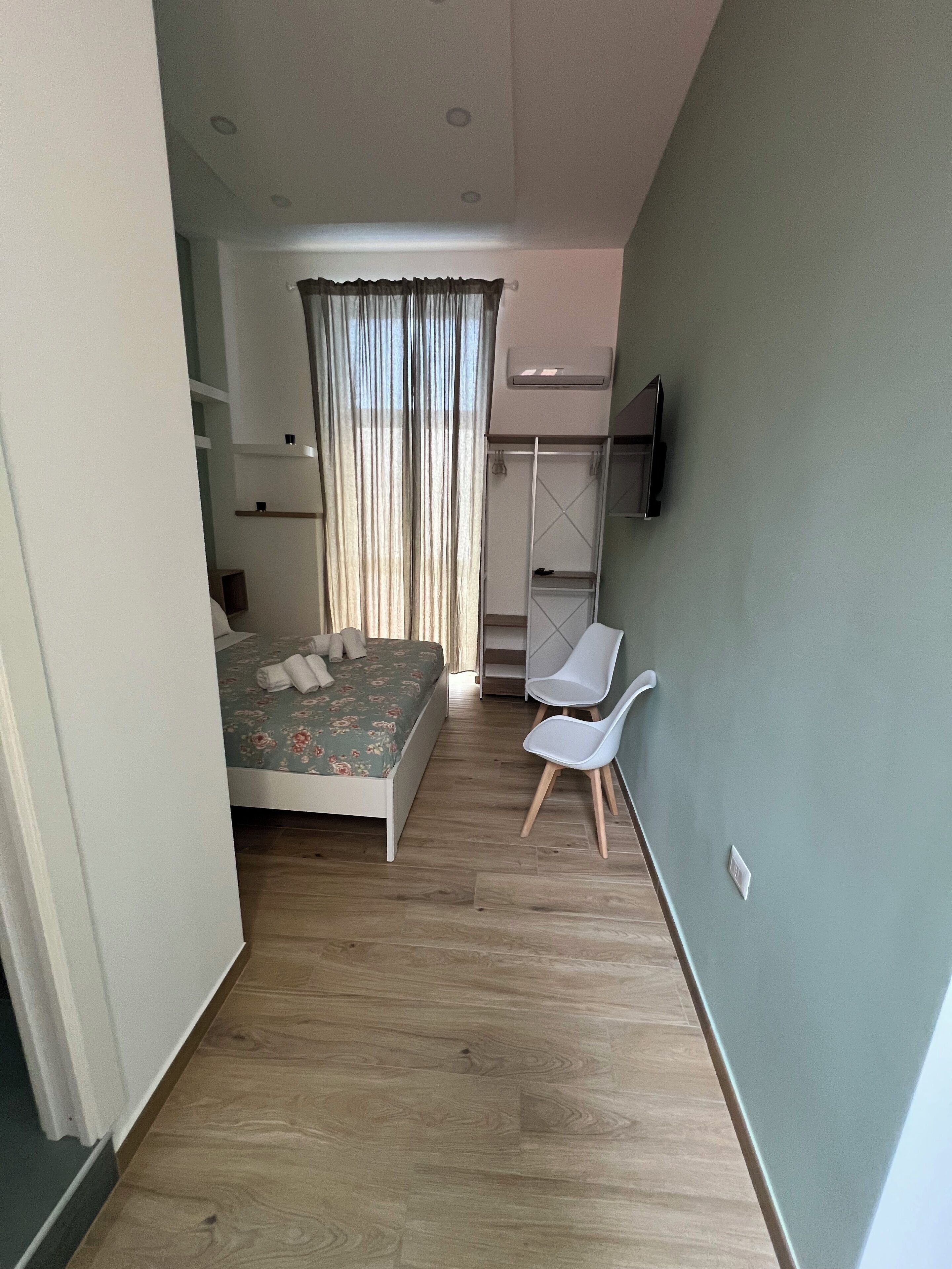 A bedroom with a bed, a desk, and a chair. There is a window with curtains, a TV mounted on the wall, and a wardrobe.