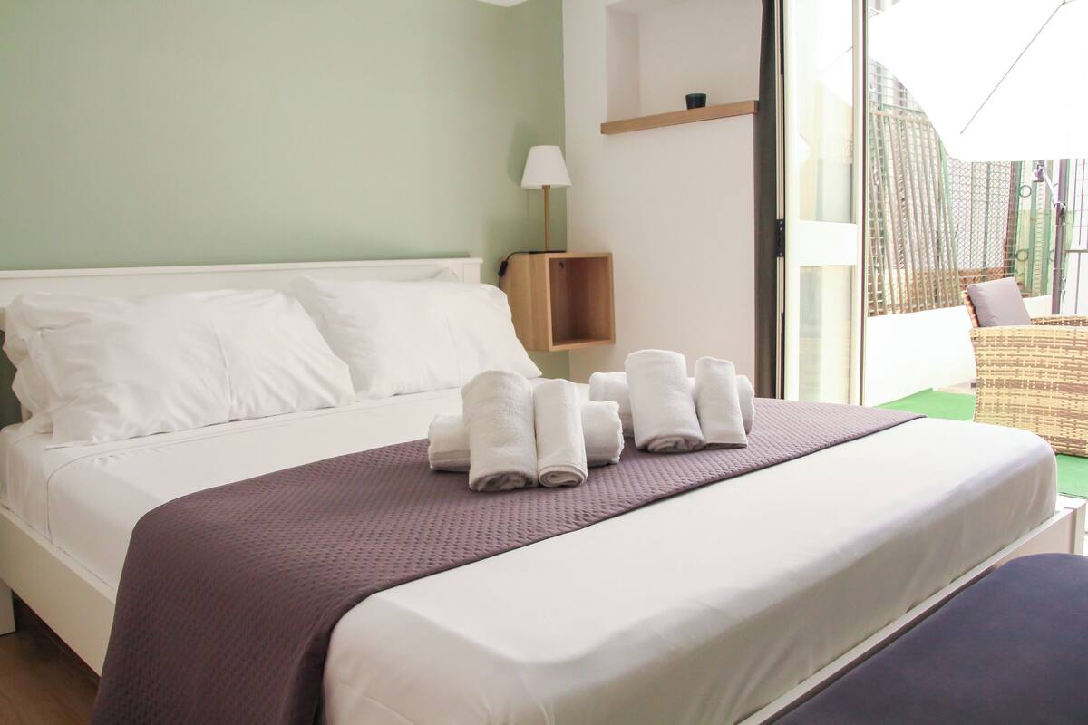 A neatly made bed with white linens and a purple blanket, a wooden nightstand with a lamp, and a view of an outdoor area through a glass door.