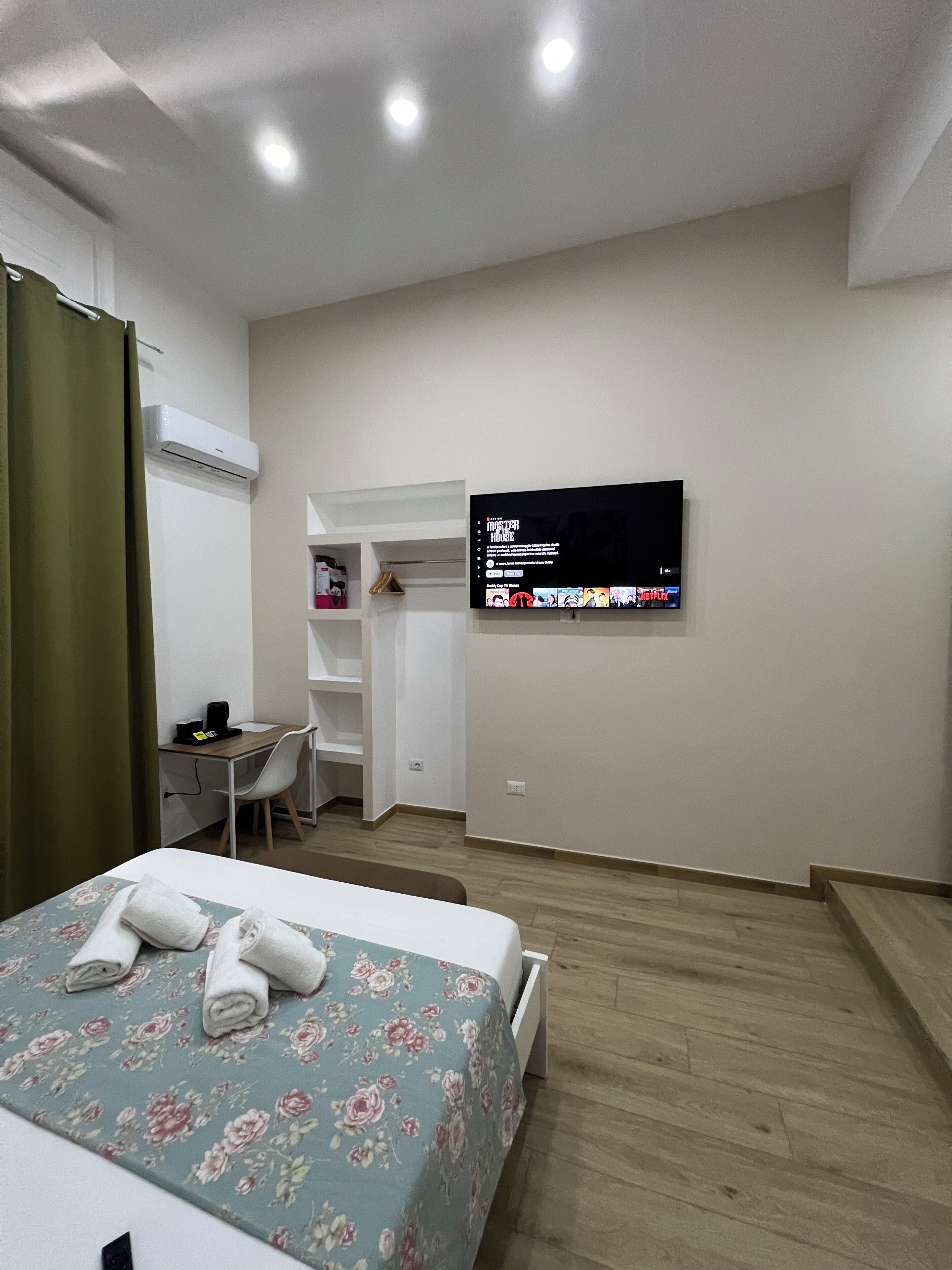A hotel room with a bed, a desk, a chair, a TV mounted on the wall, and an air conditioning unit.