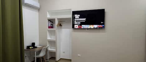 A hotel room with a bed, a desk, a chair, a TV mounted on the wall, and an air conditioning unit.