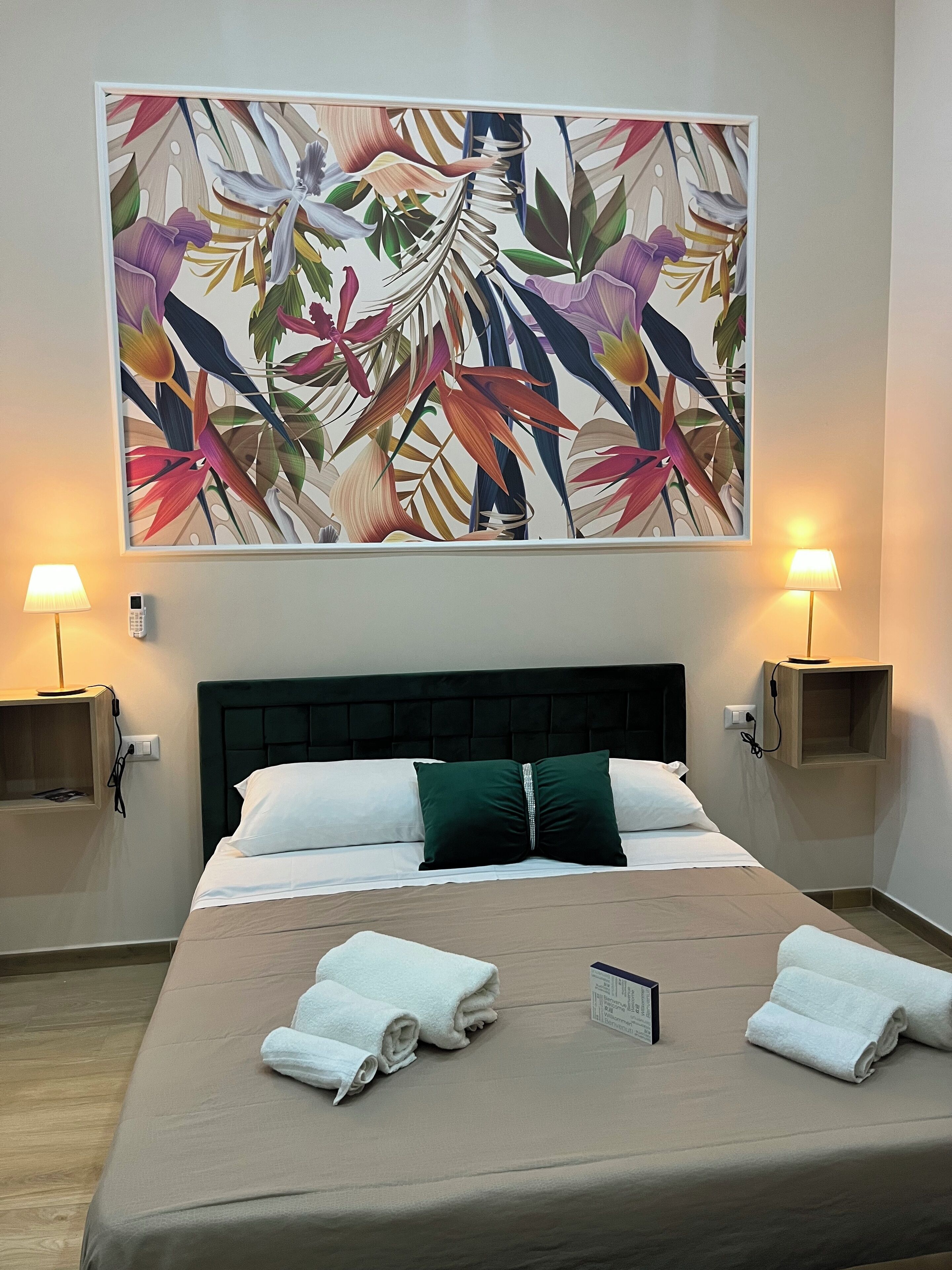 A hotel room with a large floral artwork on the wall, a bed with white and green pillows, two bedside lamps, and neatly folded towels on the bed.