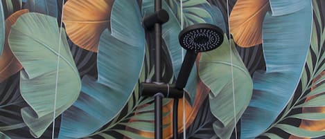 A shower with a black head and a handheld brush, set against a wall with a large tropical leaf pattern.