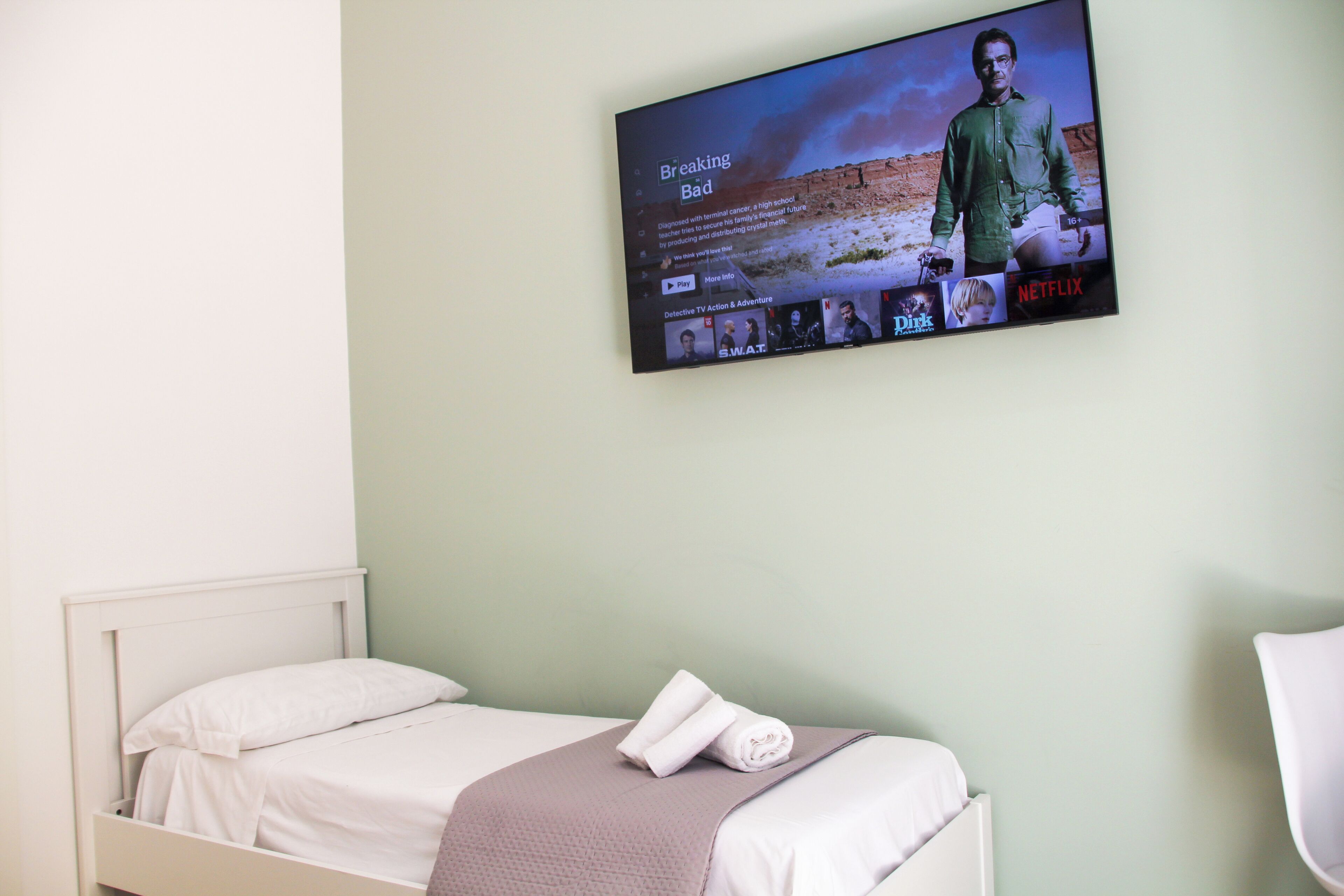 A hotel room with a single bed, a TV mounted on the wall displaying a movie, and a neatly made bed with a towel.